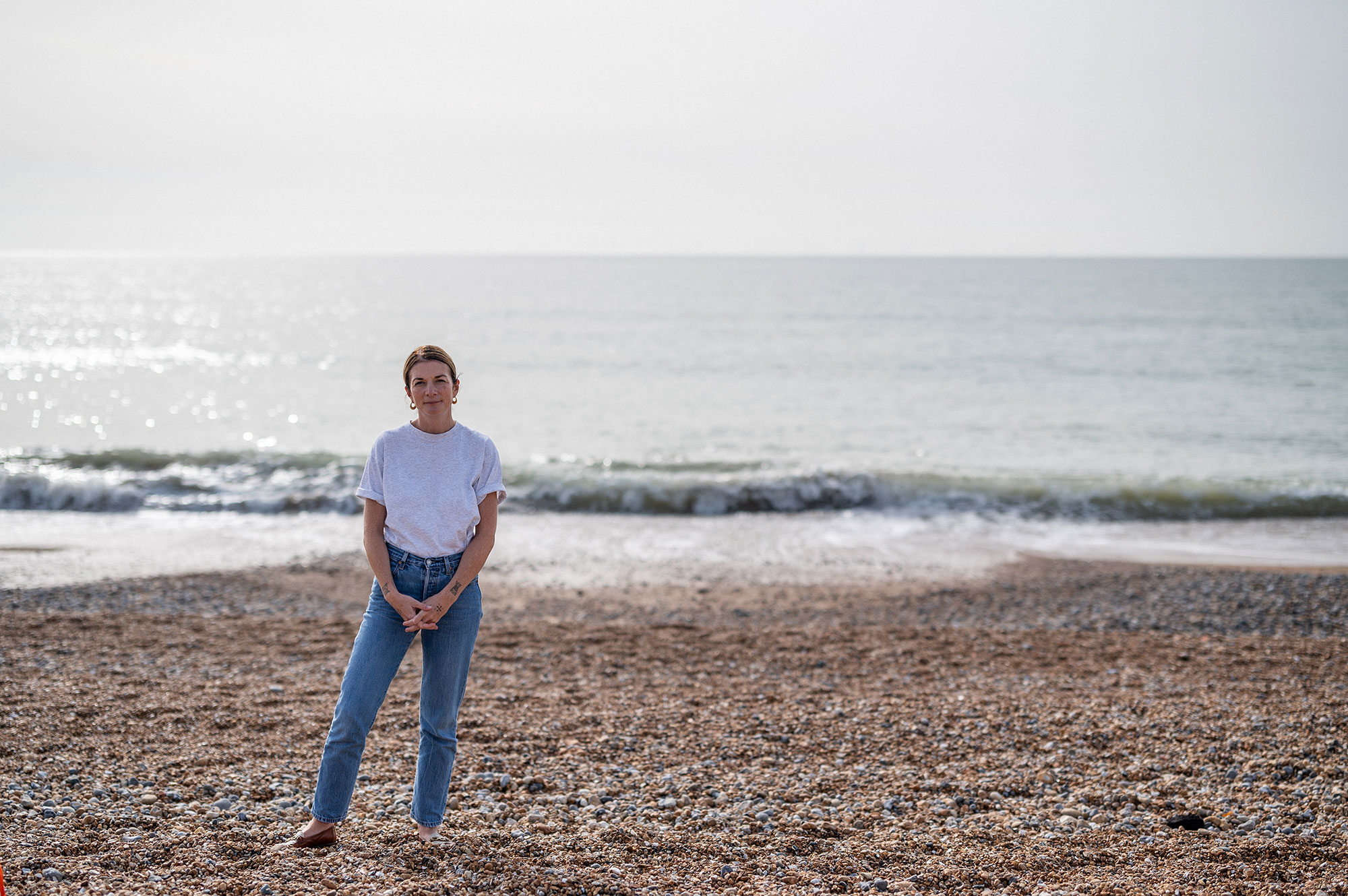 Eliza Fricker stands on the beach at Brighton facing the camera with the sea behind her